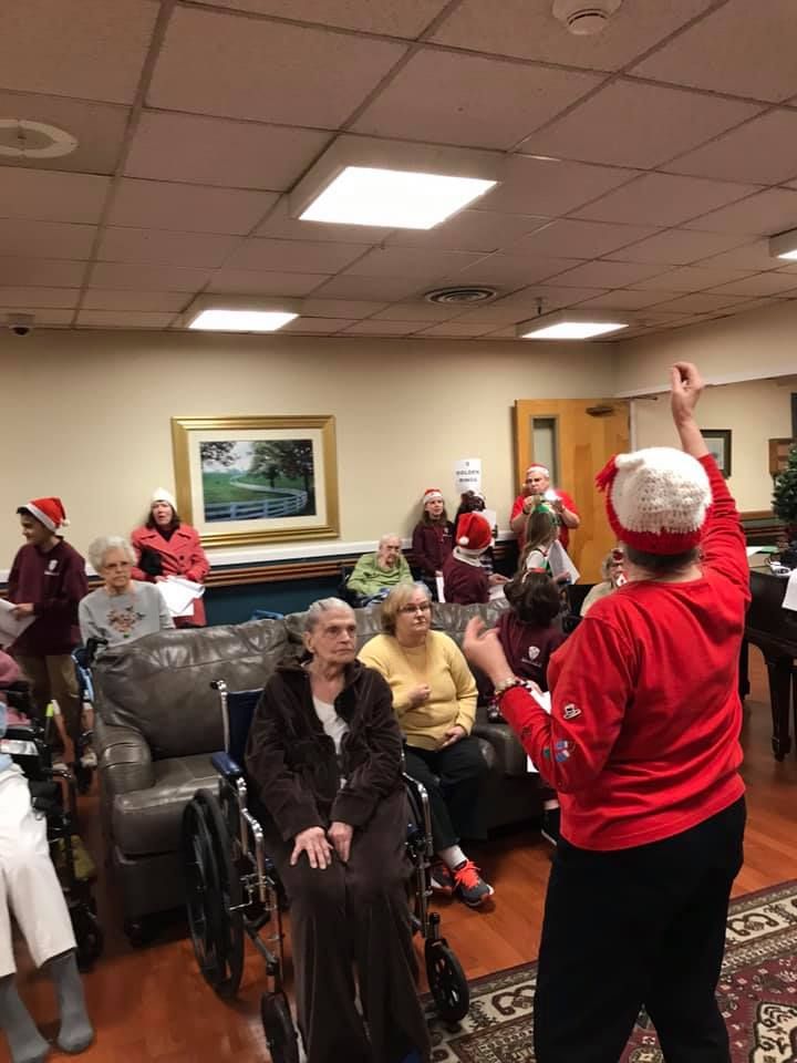 Group of people singing, some wearing Santa hats, in a room. One person conducts.