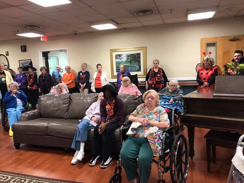 Group of seniors in a room, some seated, some standing, looking towards the right, near a piano.