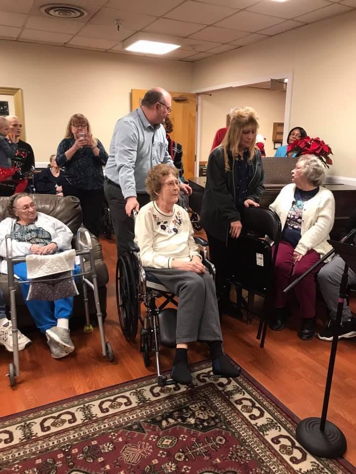 People in a room, some in wheelchairs. A man leans to talk to a woman in a wheelchair, another woman stands nearby.