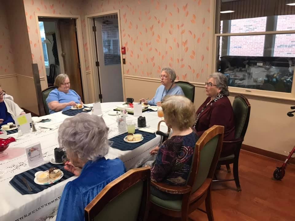 People seated at a table, eating and conversing in a dining area. Some wear blue tops. A small red cart is visible.