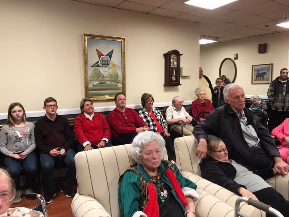 People sitting in a room, some in red attire. A clock and framed art hang on the wall.