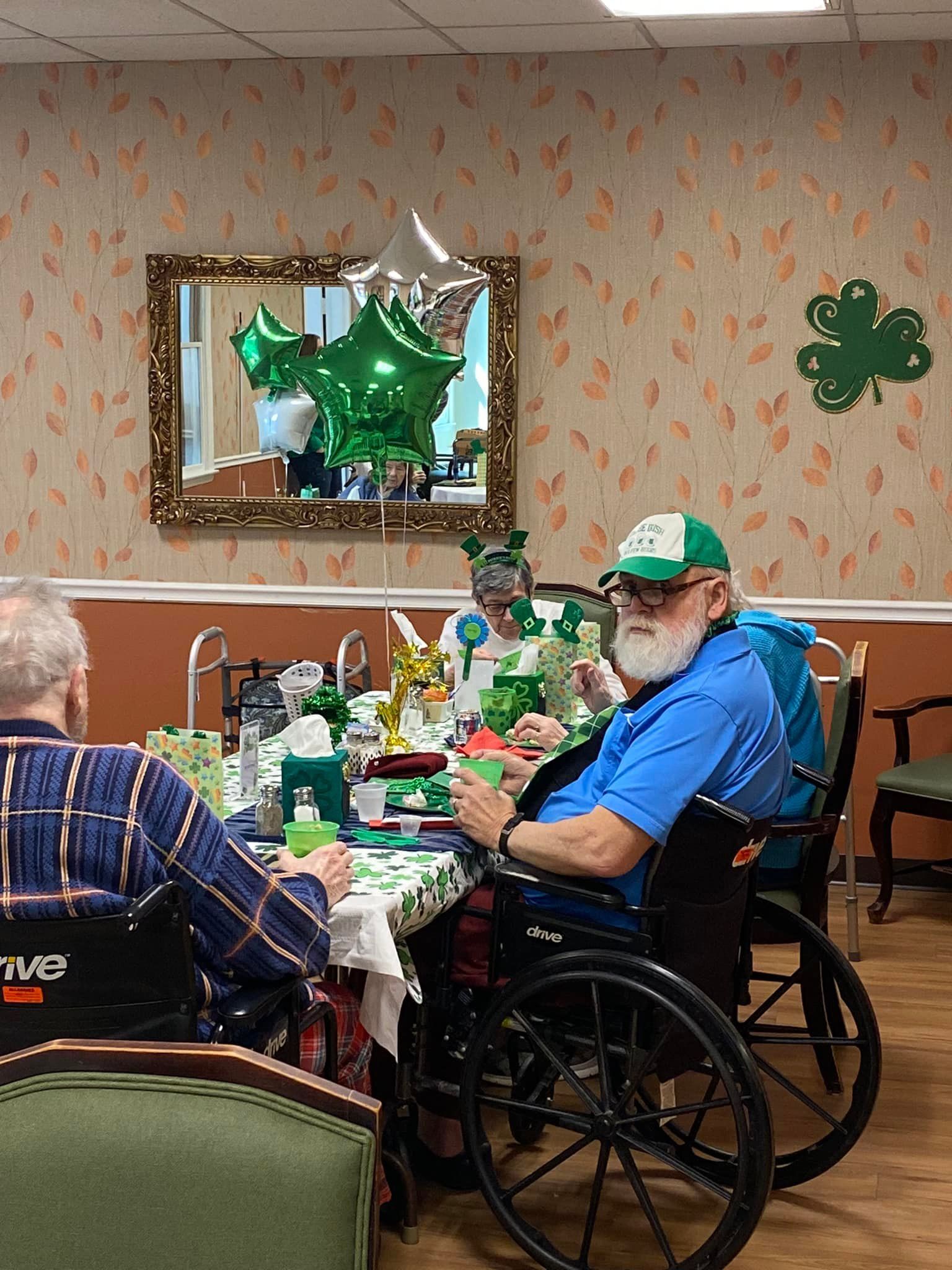 People in wheelchairs celebrate St. Patrick's Day at a table with decorations, balloons, and a mirror.