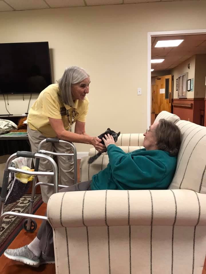 Woman in yellow shirt hands a small black cat to a seated woman in a teal sweater, near a walker.