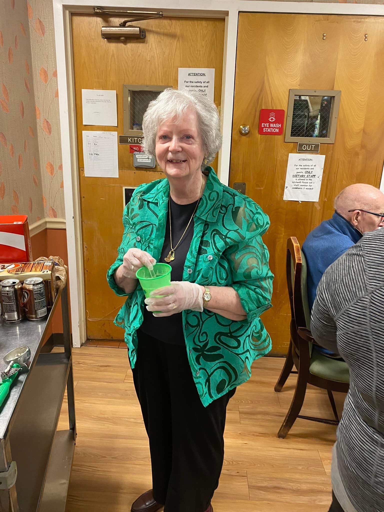 Woman in green jacket holds a cup, smiling, near a food prep area.