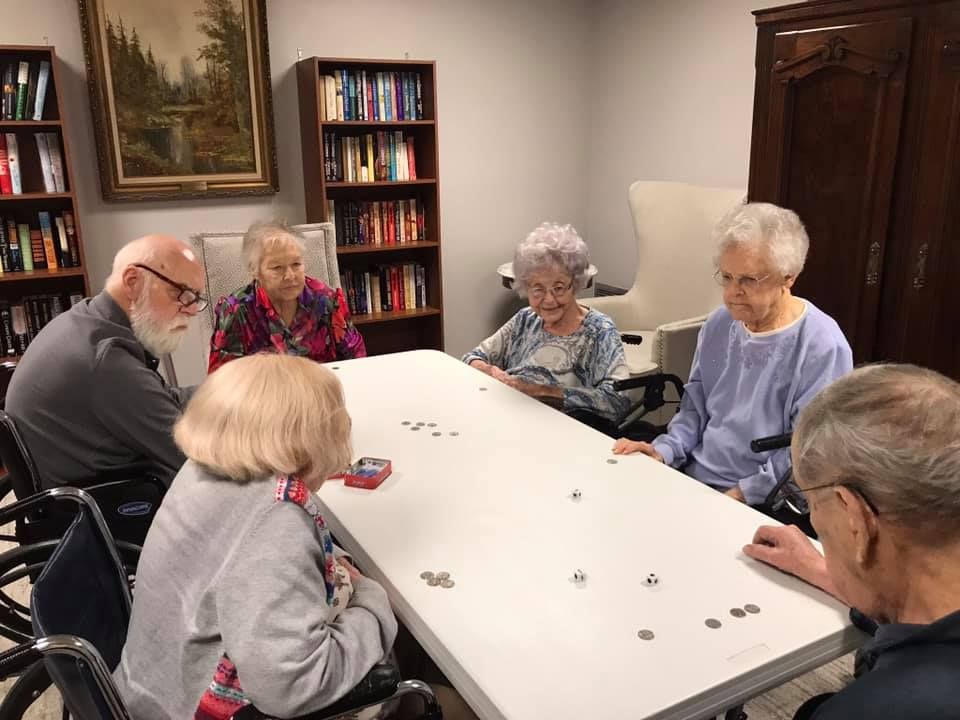 Seniors playing a board game at a table in a library with books and art.