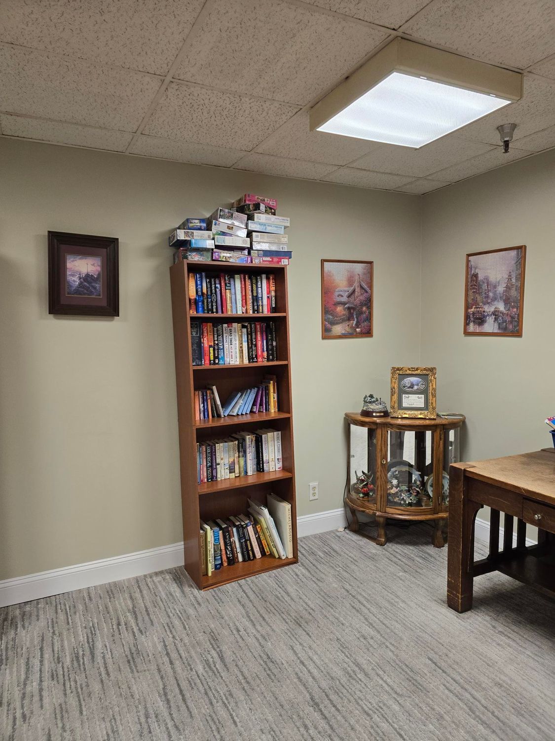 A room with a bookcase, desk, and framed pictures. Books are stacked on top of the bookcase.