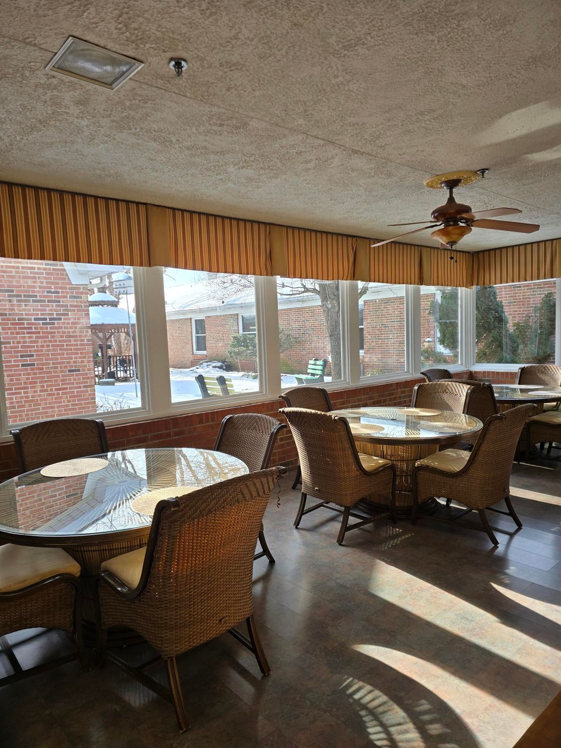 Sunroom with wicker furniture, round tables, and large windows overlooking a backyard.