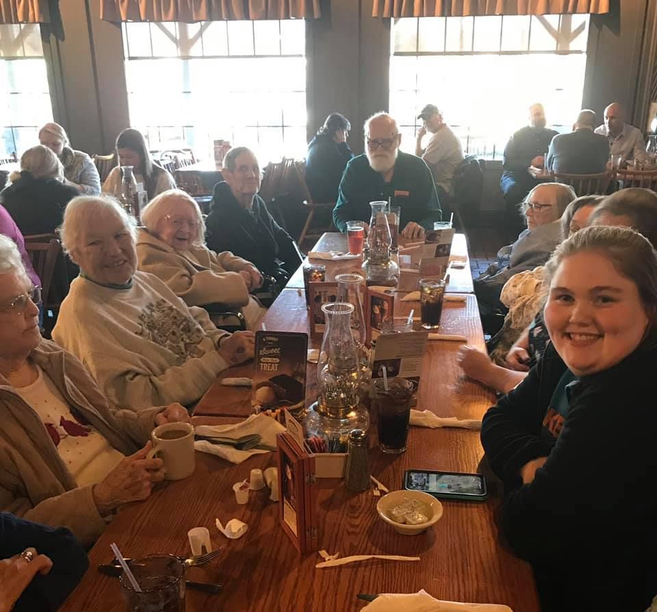 People seated at a long table in a restaurant; smiling, drinking, and looking at the camera. Sunlight through windows.