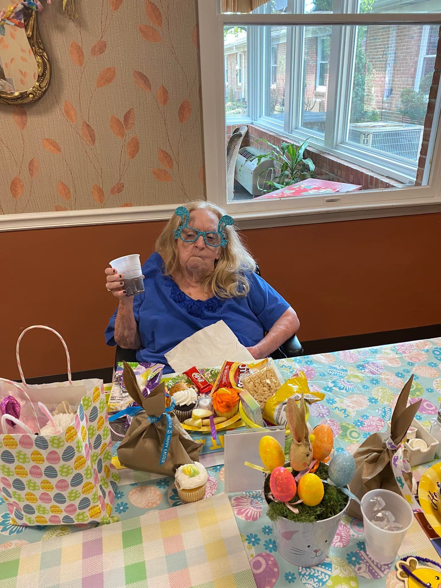 Woman holding a cup at a decorated Easter table. Bunnies, eggs, cupcakes, and gift bags are visible.