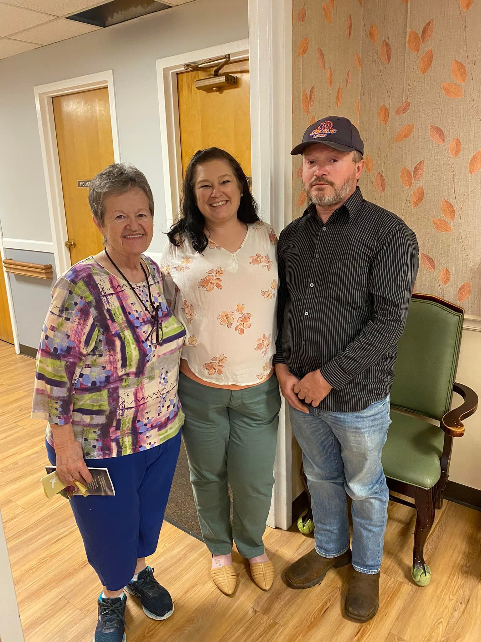 Three people standing in a room with light wood floors and wallpaper. The woman in the middle is wearing a white floral shirt.