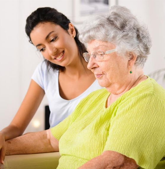 Woman in white top looking at elderly woman in green shirt; indoors.