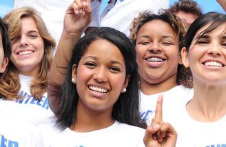 Group of smiling people wearing white shirts, raising hands, one pointing up.