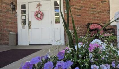 White door with a pink wreath and flowers in front of a brick building.