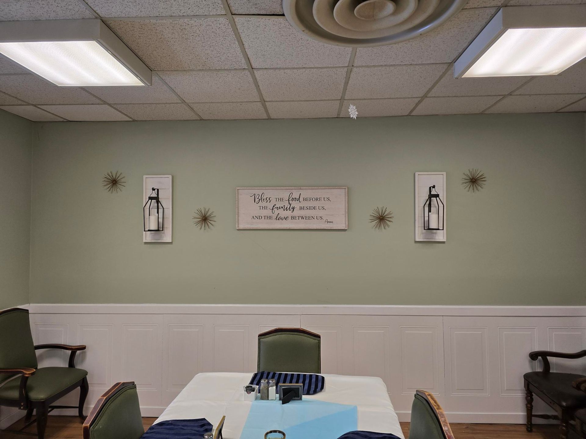 Dining room with a light green wall, white wainscoting, and a white tablecloth. Decorative artwork and chairs surround the table.