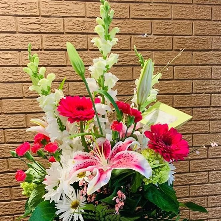 Floral arrangement with pink lilies, red gerbera daisies, and white snapdragons against a brick wall.