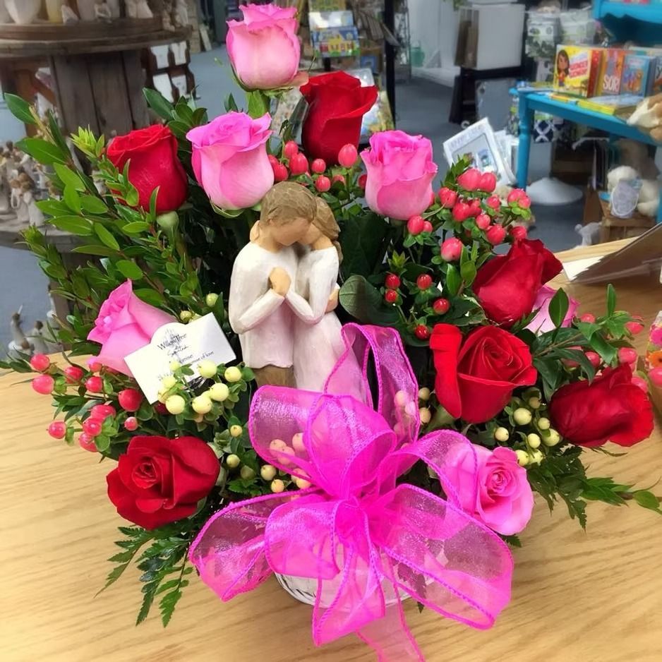 Floral arrangement with red and pink roses, greenery, a figurine, and a pink bow.
