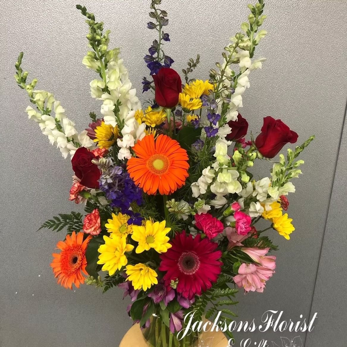 Colorful flower arrangement in a vase, including red roses, orange and purple gerbera daisies, and white snapdragons.
