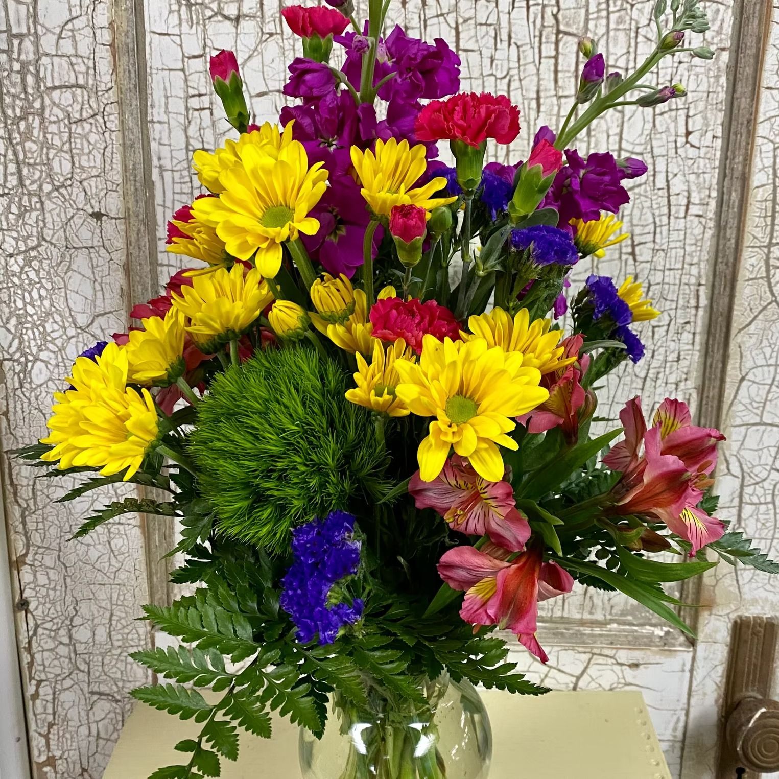 Vase of vibrant flowers, including yellow daisies, pink alstroemeria, purple carnations, and blue statice.