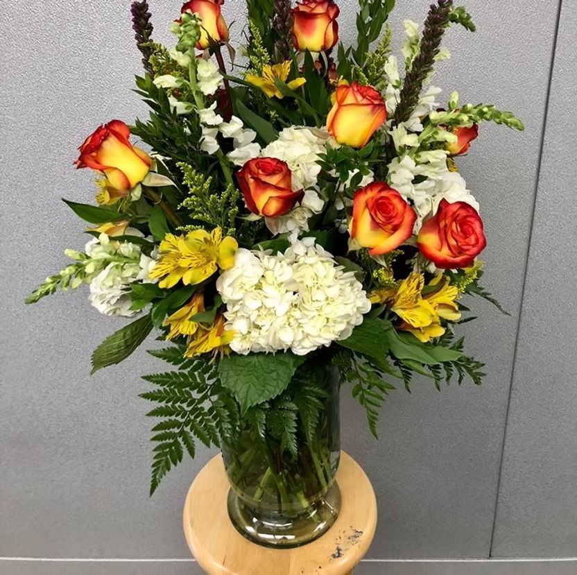 Bouquet of orange roses, white hydrangeas, yellow flowers, and greenery in a glass vase.