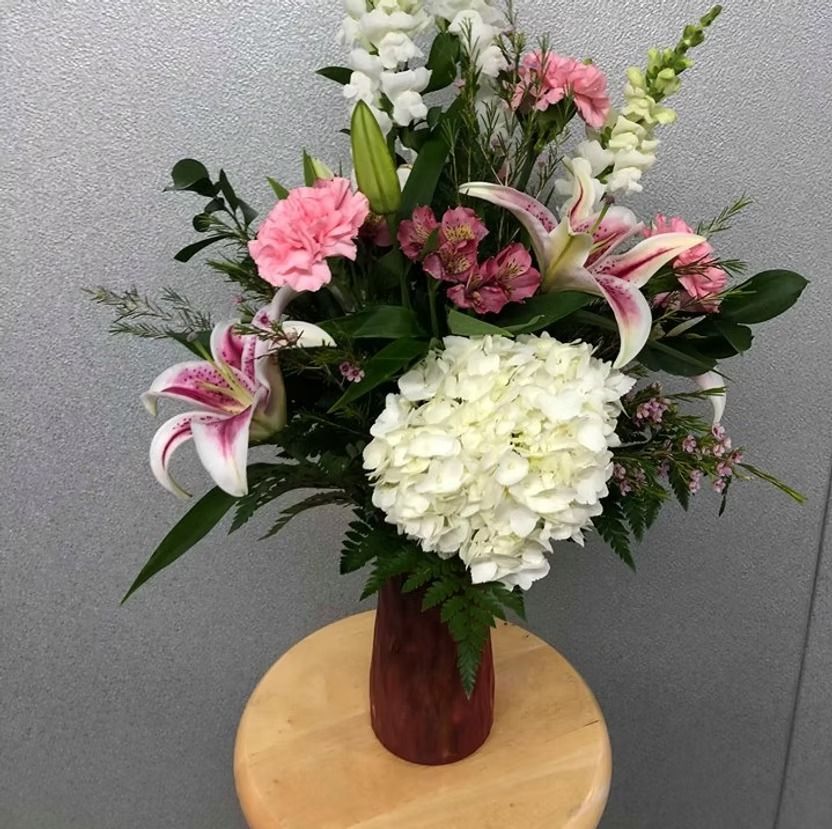 A floral arrangement with pink and white lilies, carnations, and hydrangea in a maroon vase on a wooden stool.