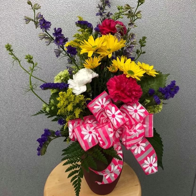 Arrangement of yellow, white, red, and purple flowers with a pink daisy ribbon, in a brown vase.