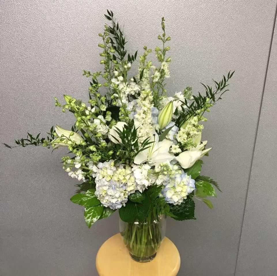 White and light blue flower bouquet in a clear vase on a small wooden stand, against a light gray wall.