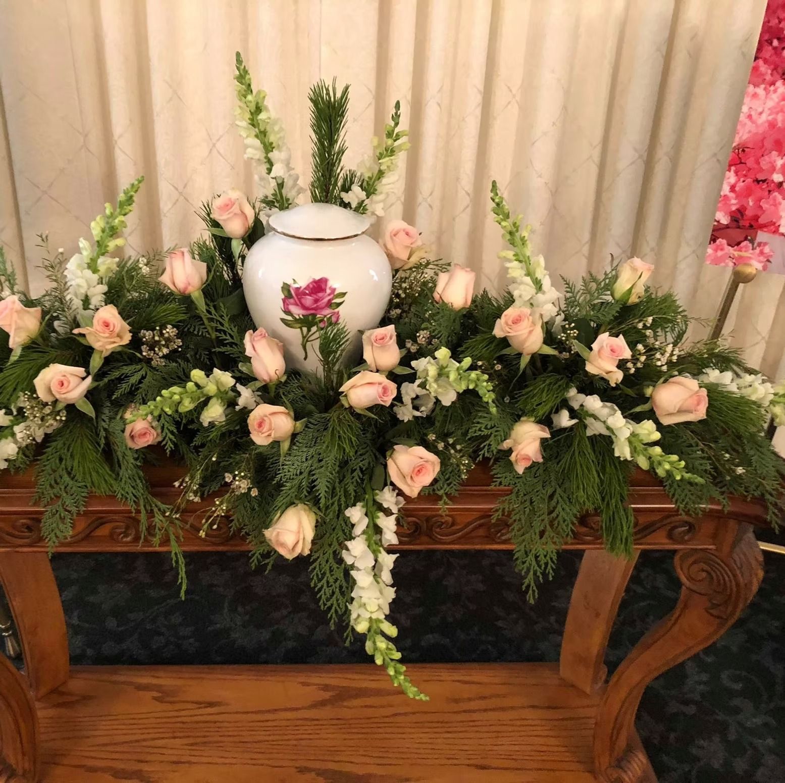 Urn surrounded by floral arrangement with pink roses and white flowers on a wooden table.