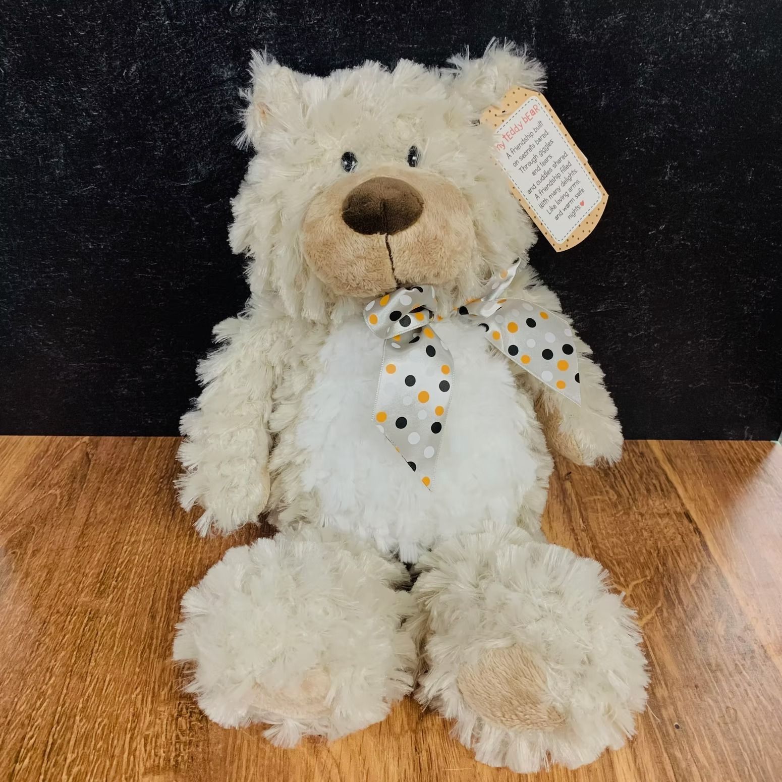 Beige plush teddy bear with a polka-dot bow tie. The bear has a brown nose and a tag.