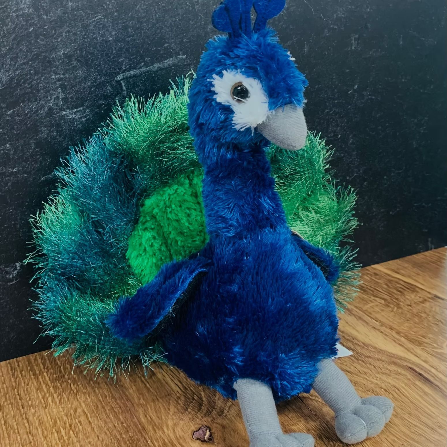 Stuffed peacock toy, blue body, green and blue tail feathers, perched on a wooden surface.