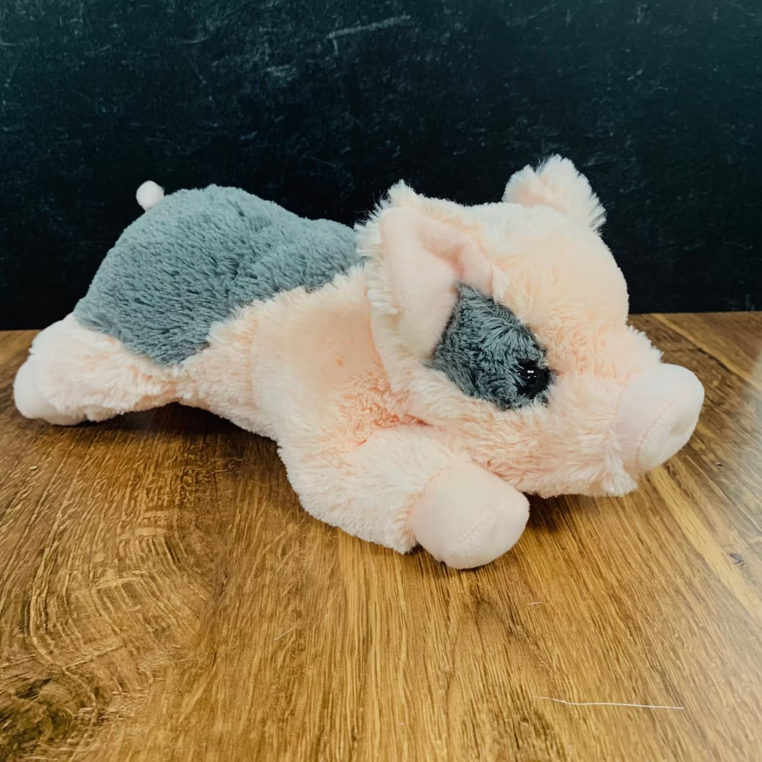 Plush pig with pink body, gray spot on eye and back, lying on a wooden surface.