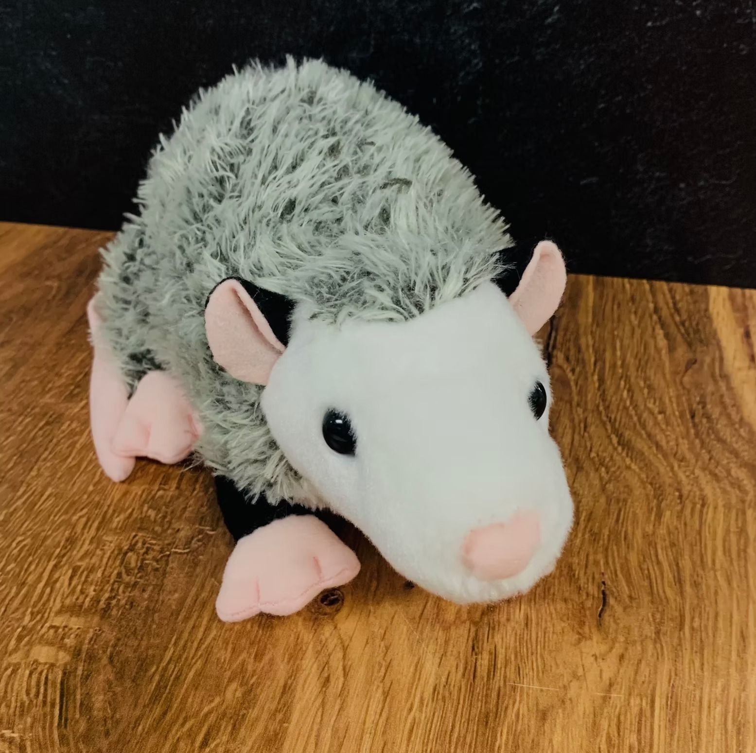 Stuffed opossum with gray fur, pink ears, and a white face on a wooden surface.