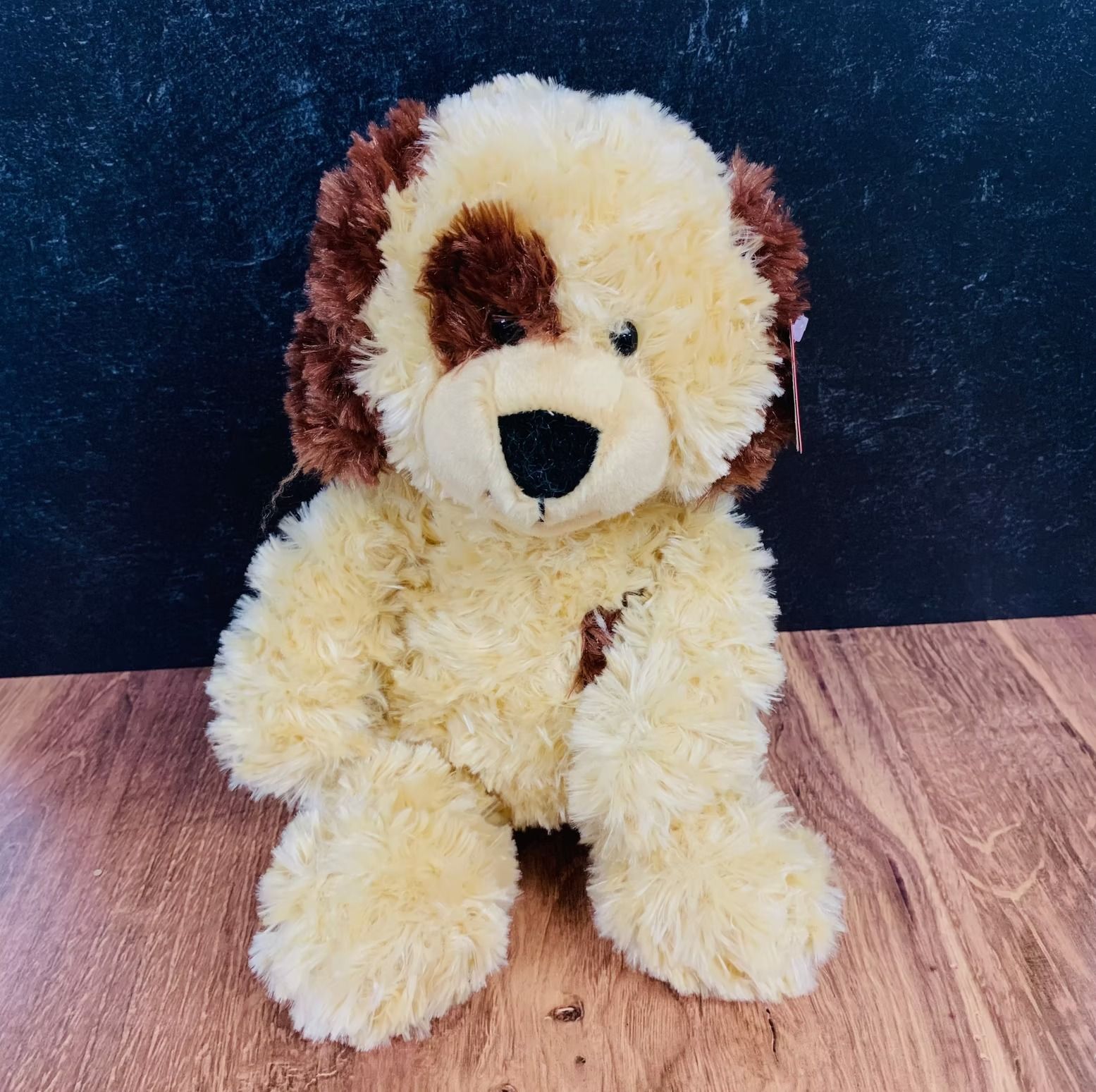 Stuffed dog toy with tan and brown fur, sitting on a wooden surface in front of a dark background.