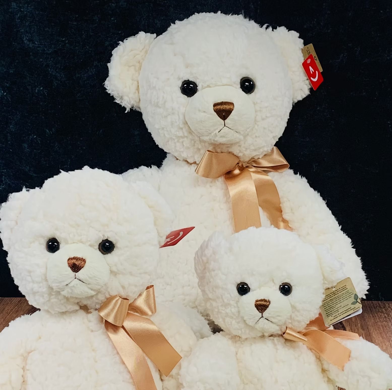 Three cream-colored teddy bears: large, medium, and small, with brown eyes and ribbon bows, against a dark backdrop.