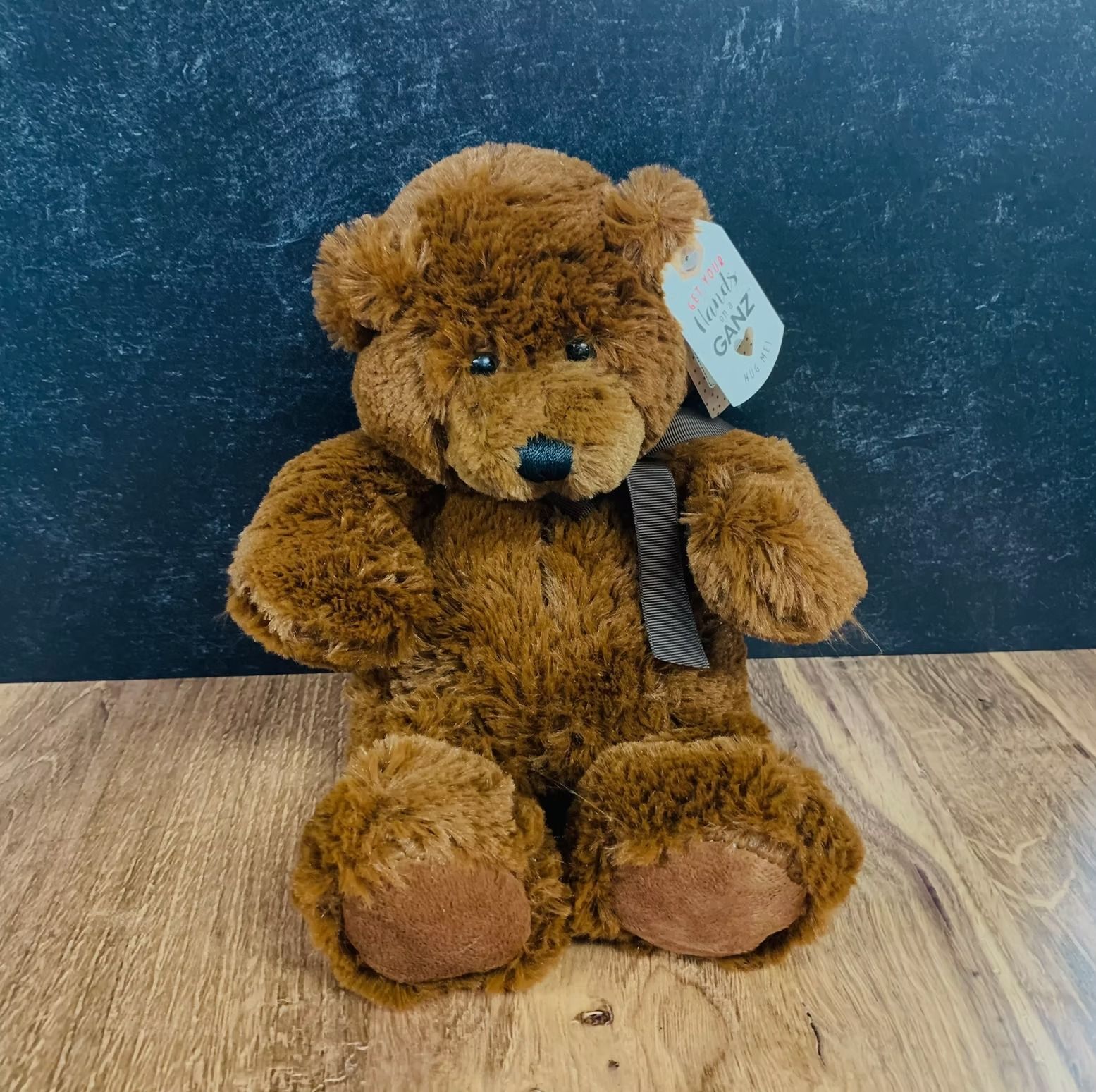 Brown teddy bear with a black scarf, sitting on wood. It has a tag.