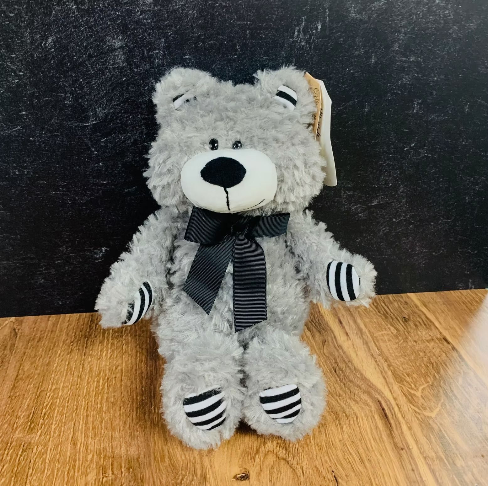 Gray teddy bear with a black bow tie and striped accents, set on wood against a dark backdrop.