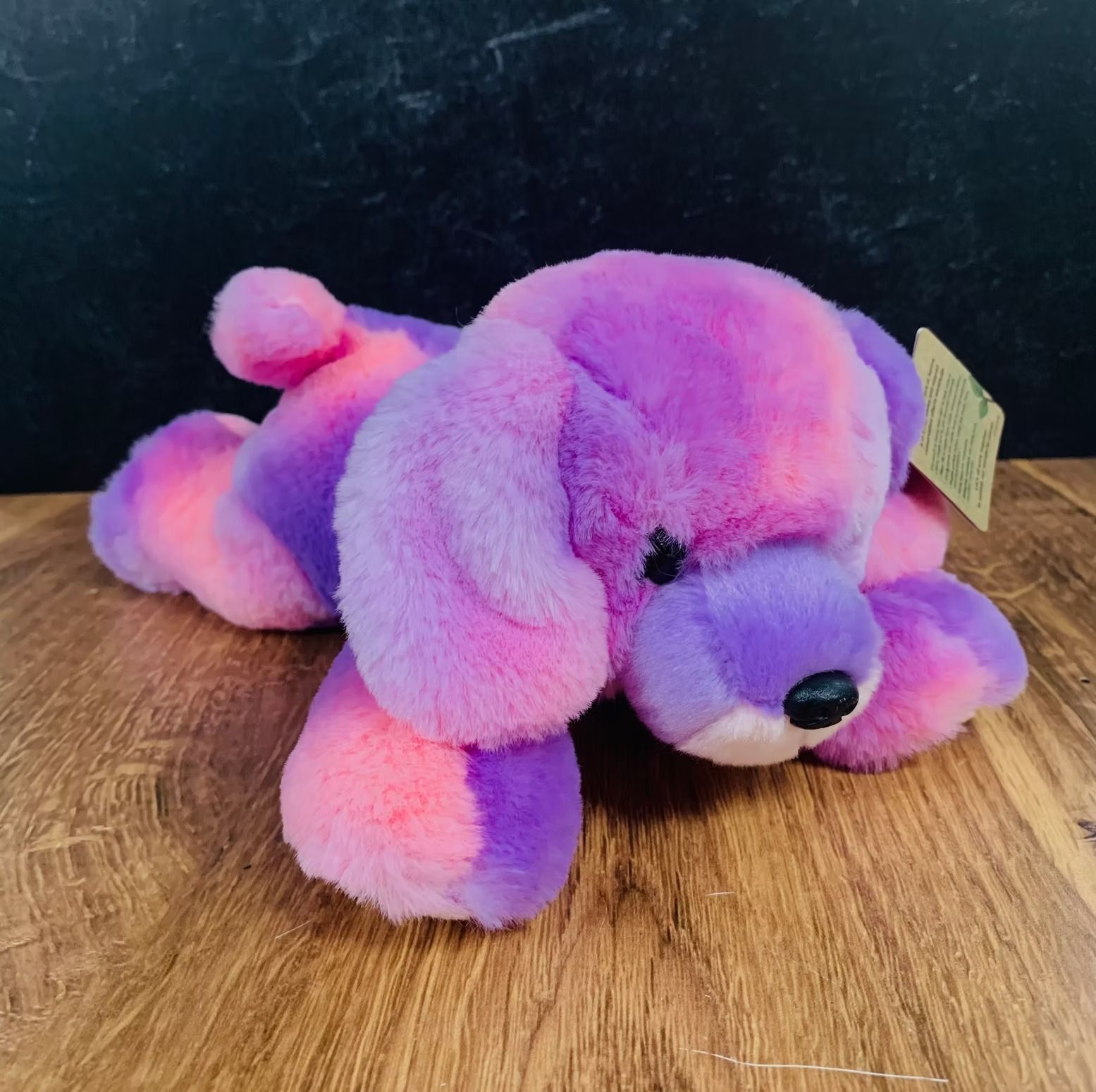 Plush dog toy, pastel pink and purple colors, lying on wood surface. Black eyes and nose, tag attached.