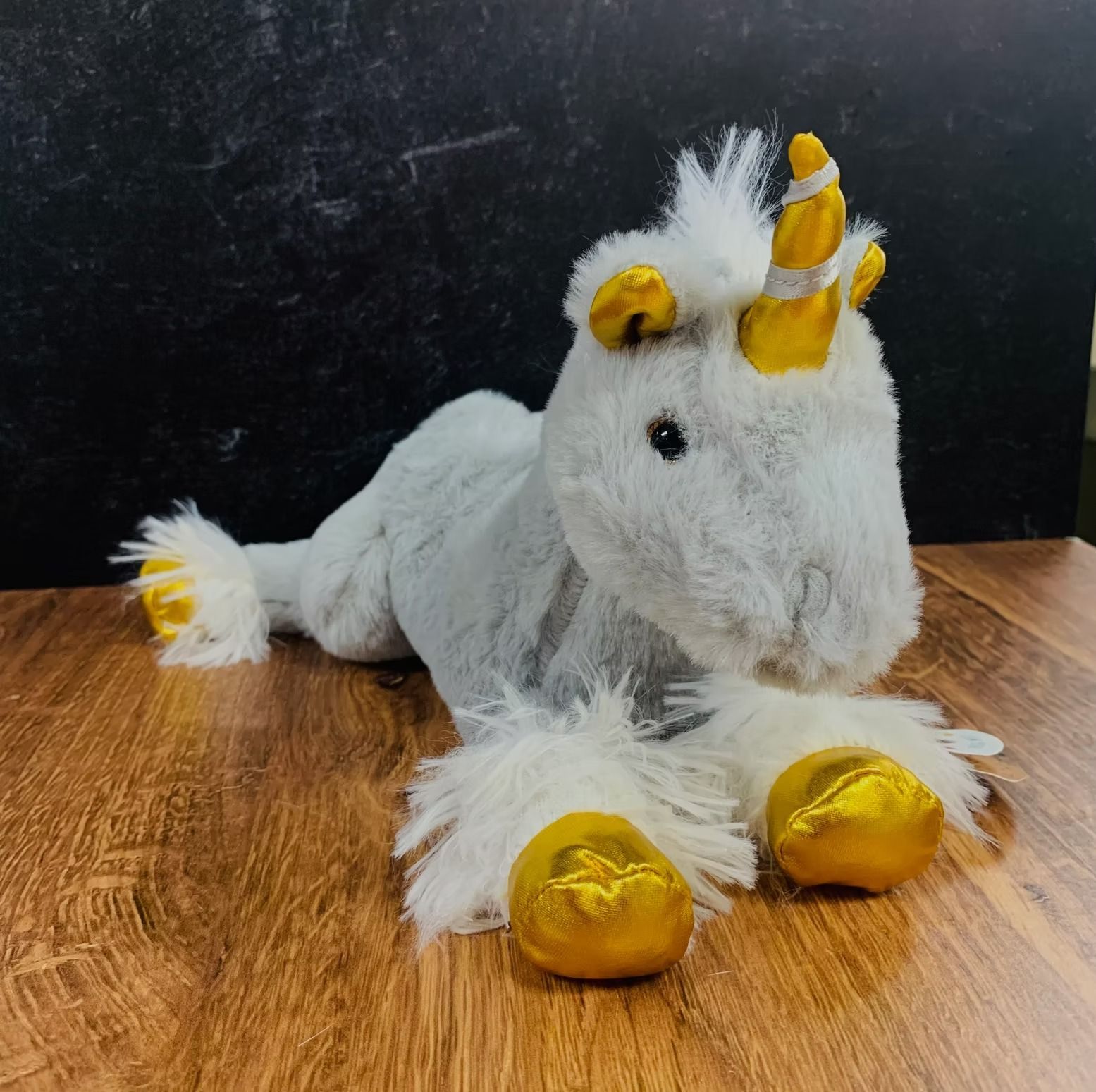 Fluffy, plush unicorn toy with golden hooves and horn, lying on a wooden surface.