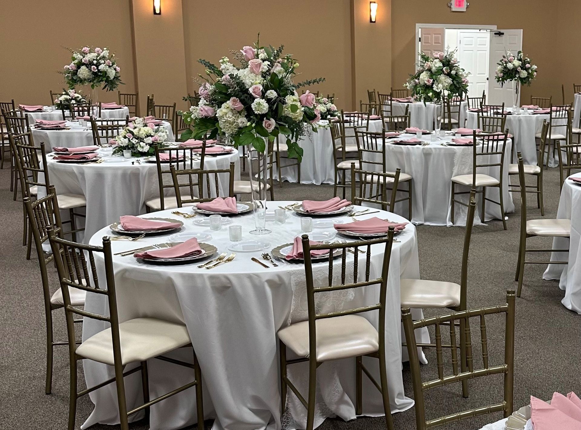Reception hall with round tables set for a formal event. White linens, floral centerpieces, gold chairs, and pink accents.