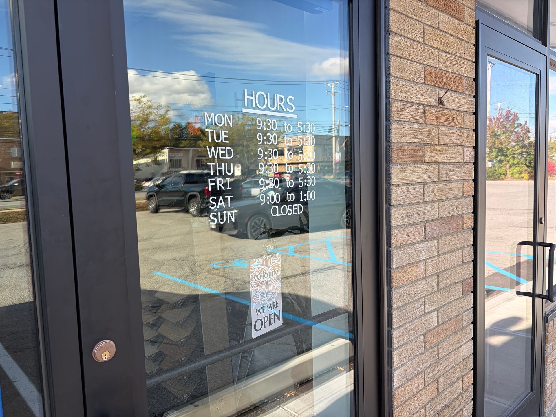Exterior of a business with hours posted on the glass door. Brick wall to the right. Car and blue sky reflected.