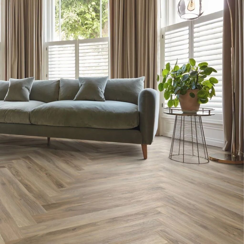 Living room with a gray sofa, a patterned wooden floor, and a plant near a window with shutters.
