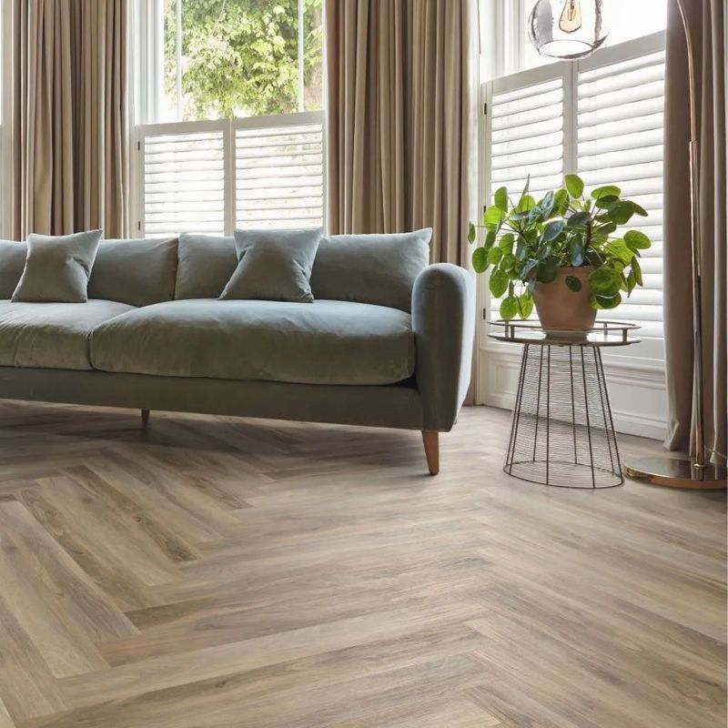 Living room with a gray sofa, a patterned wooden floor, and a plant near a window with shutters.