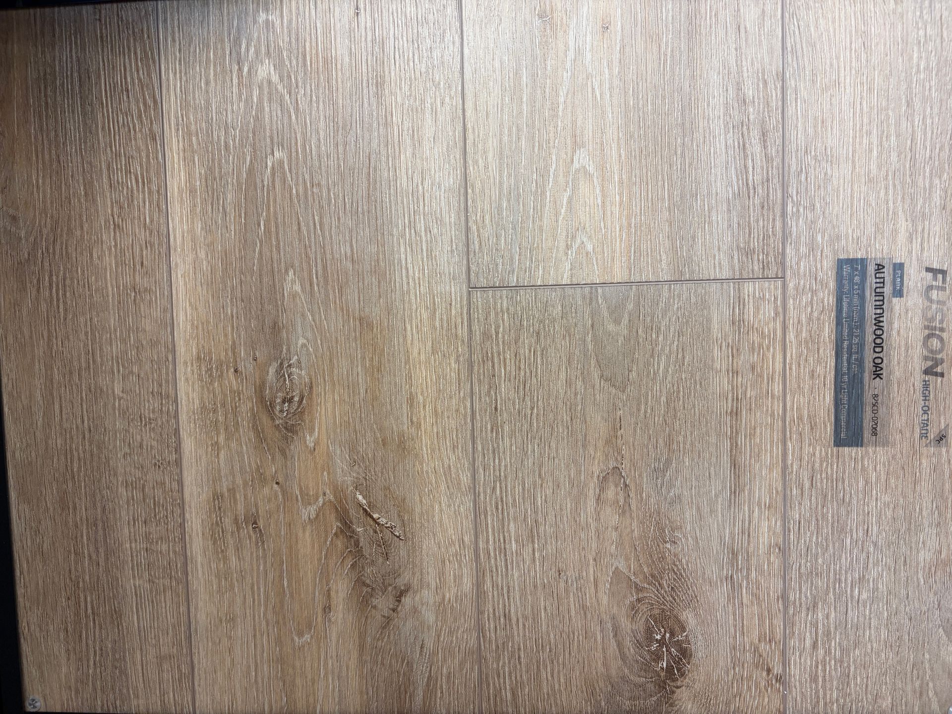 Wood-look flooring with a visible crack; the surface is light brown with darker knots and a label in the corner.