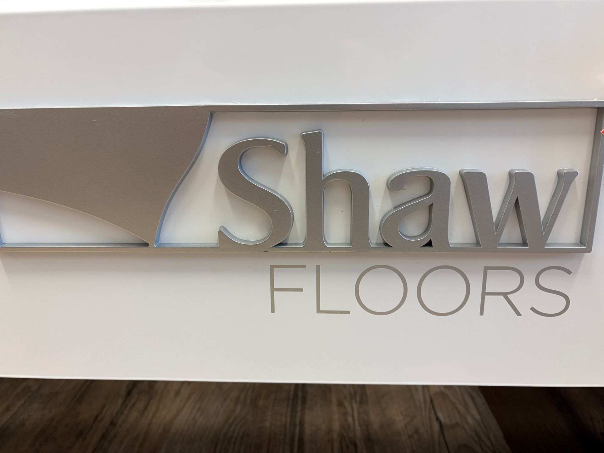 Shaw Floors logo on white surface with a silver metallic accent. 