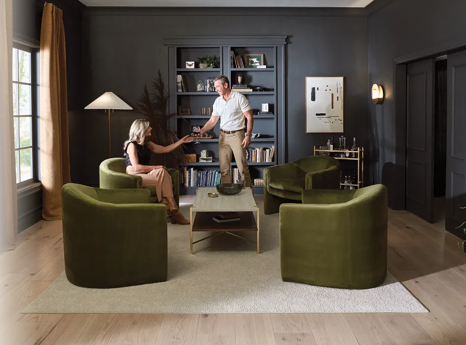 A couple toasting in a dark-walled living room with olive green armchairs and a built-in bookcase.