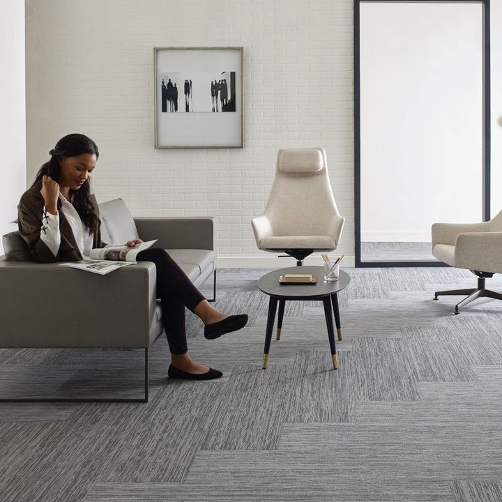 Woman reading on a sofa in a modern office waiting area. Grey carpet, white walls, and a black framed art piece.
