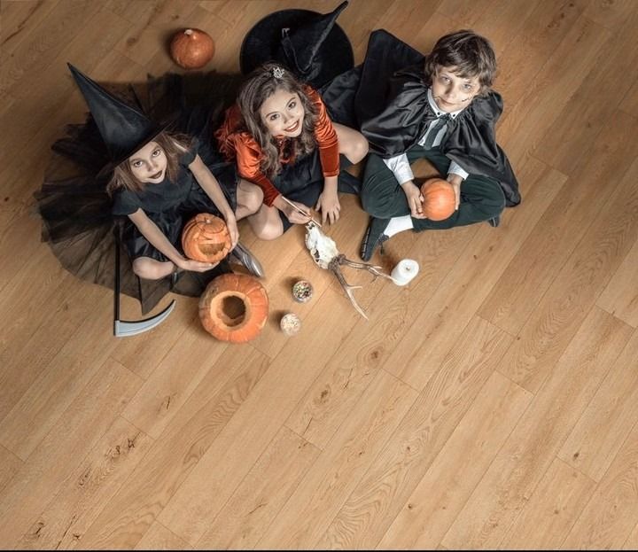 Three children in Halloween costumes sit on a wood floor with pumpkins, a skeleton, and a witch hat.