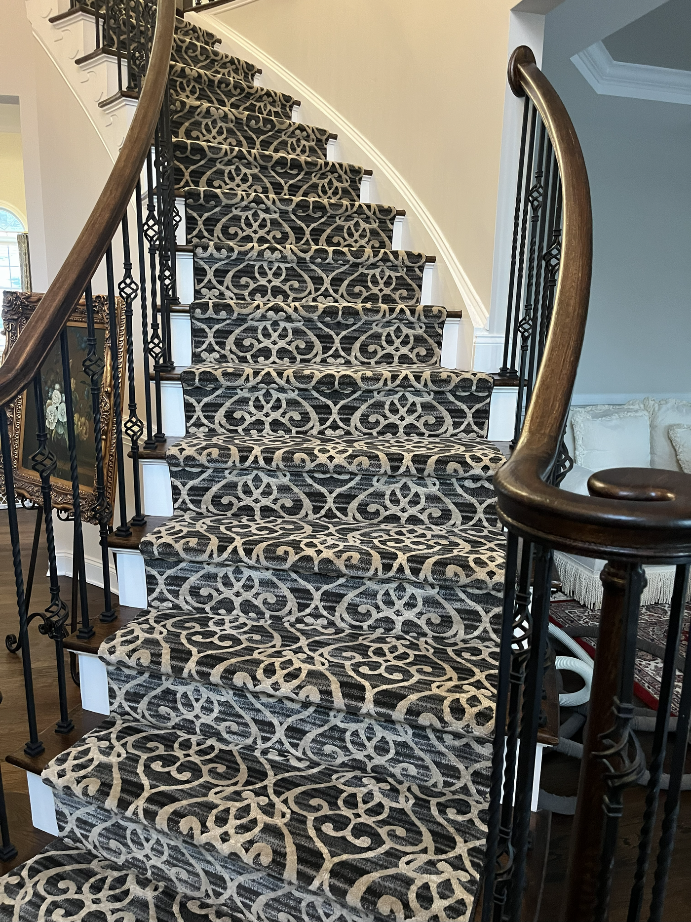 Patterned carpet covers curved staircase. Dark wood banister and ornate iron railing. White risers.