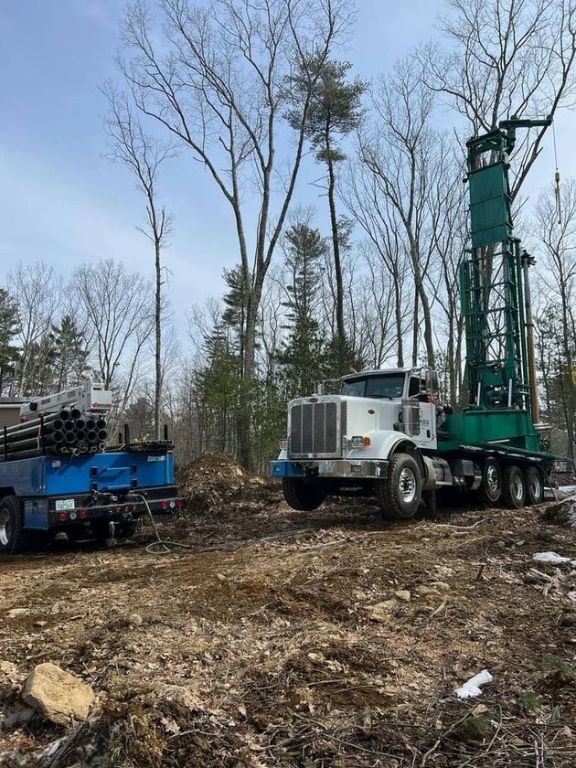 White drilling rig and blue truck in a wooded clearing with bare trees.