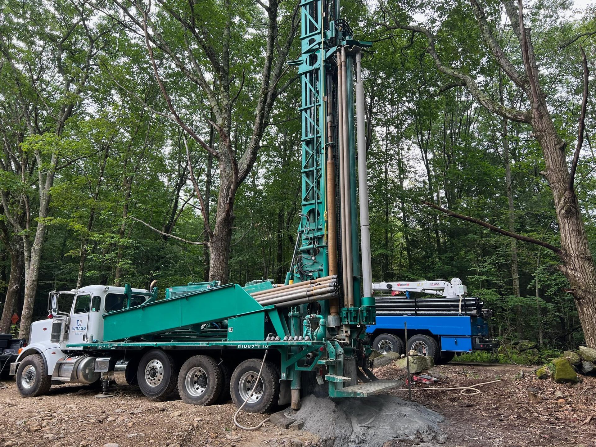Drilling rig truck in a wooded area, with a tall teal mast beside a blue truck.