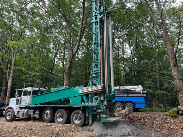 Drilling rig truck in a wooded area, with a tall teal mast beside a blue truck.